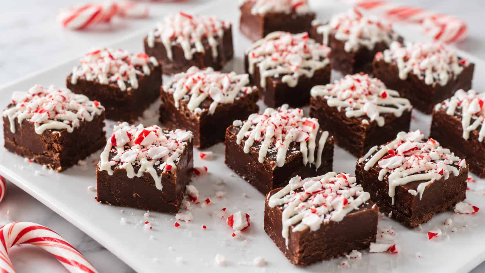 squares of chocolate fudge topped with white drizzle and crushed peppermint spread wide on a white plate, candy cane pieces scattered around, bright natural light, shallow depth of field, landscape orientation