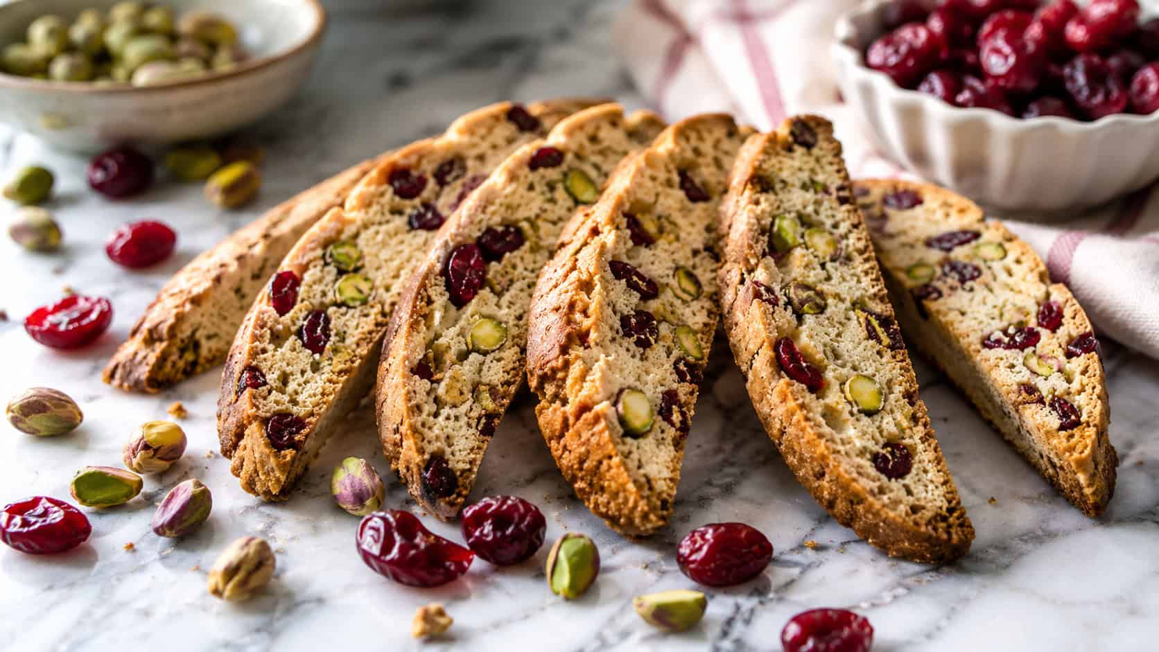 cranberry pistachio biscotti fanned wide across a marble counter, scattered red cranberries and green pistachios nearby, soft natural light, shallow depth of field, horizontal landscape composition