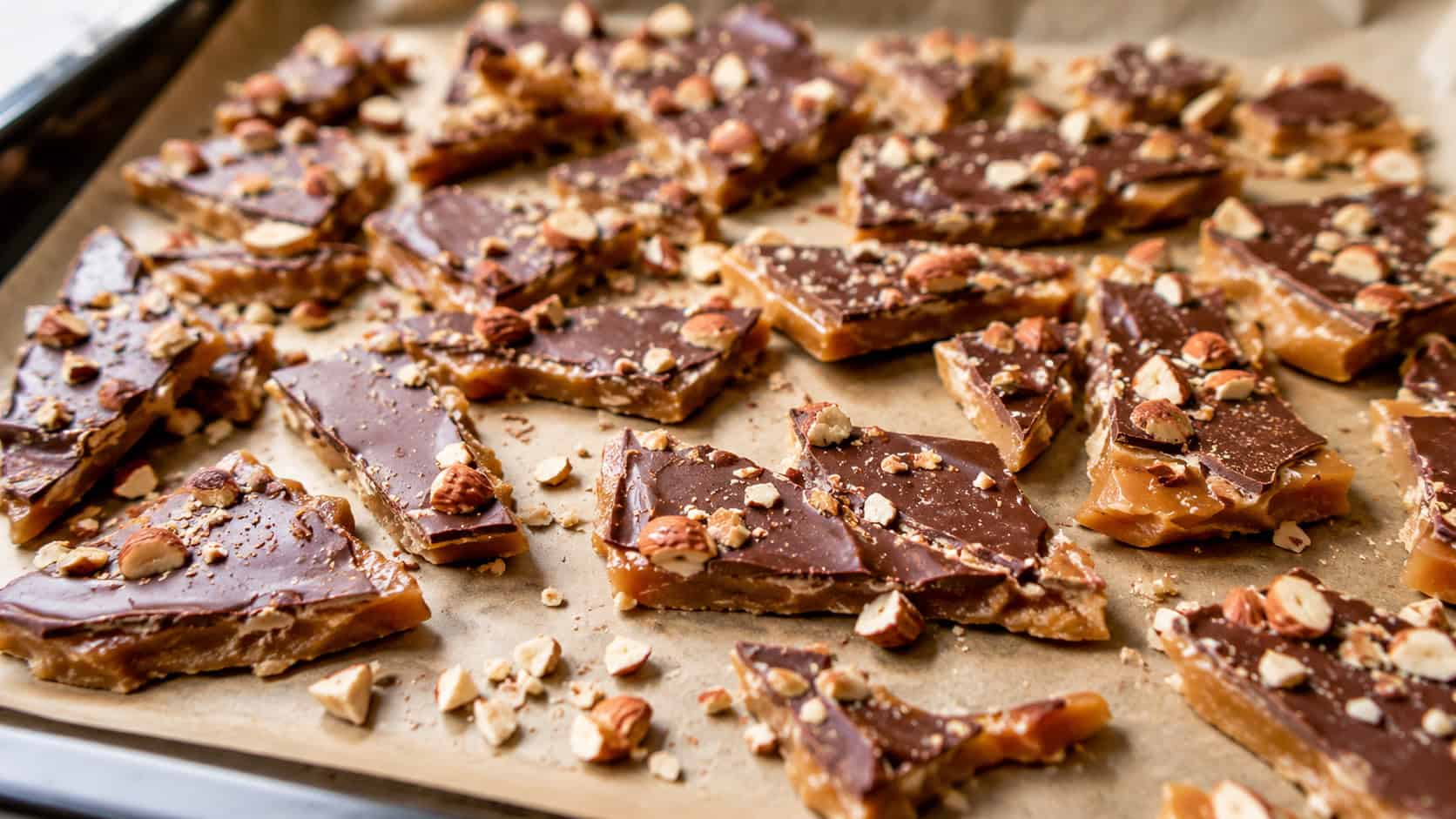 broken pieces of chocolate-topped almond toffee spread wide across a parchment-lined tray, scattered chopped nuts nearby, warm natural light, shallow depth of field, landscape orientation