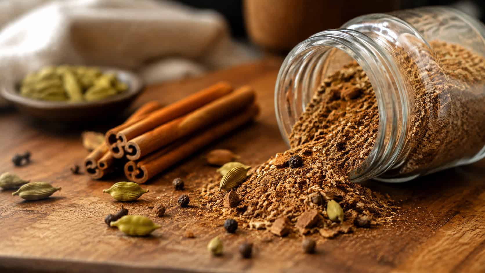 a small jar of warm-toned chai spice blend spilling slightly onto a wooden board shown wide across the frame, cinnamon sticks and cardamom pods scattered nearby, warm natural light, shallow depth of field, horizontal landscape composition