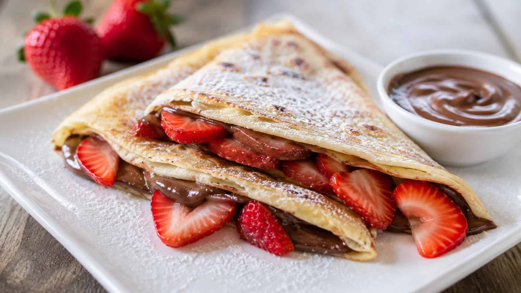 folded Nutella crepes with sliced strawberries spilling out, in a wide horizontal shot on a white plate dusted with powdered sugar, a small bowl of chocolate-hazelnut spread nearby, bright natural light, shallow depth of field, landscape orientation
