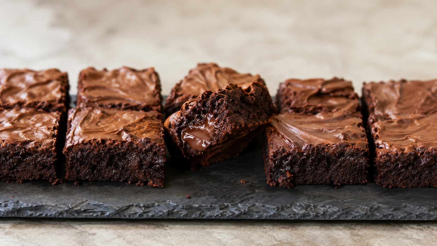 a batch of fudgy Nutella brownies with glossy swirled tops, cut into squares and arranged in a wide horizontal row on a dark slate board, one brownie broken open to show the gooey center, soft natural light, shallow depth of field, landscape orientation