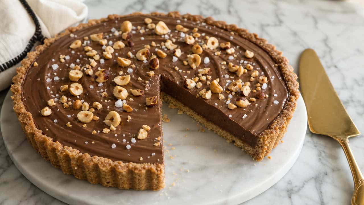 a glossy Nutella hazelnut tart topped with toasted chopped hazelnuts and flaky salt, one slice removed, in a wide horizontal shot on a marble surface with a gold serving knife, soft natural light, shallow depth of field, landscape orientation