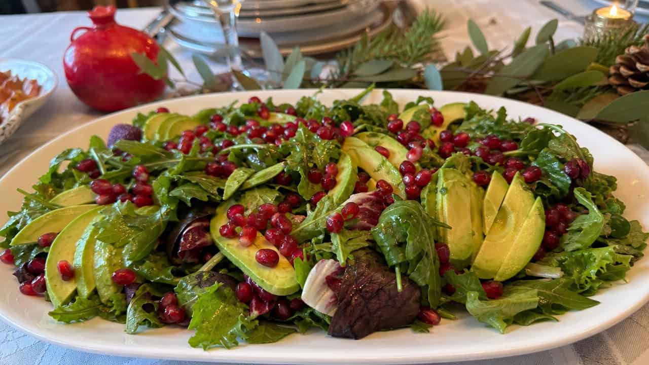 a colorful salad with sliced green avocado and ruby pomegranate arils over fresh greens in a wide horizontal shot, on a white platter on a holiday table, natural light, shallow depth of field, landscape orientation