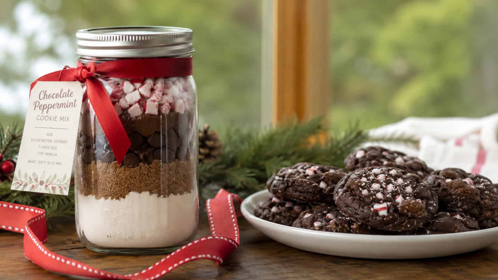 a mason jar of layered chocolate and peppermint cookie mix beside finished minty chocolate cookies in a wide horizontal shot, on a wooden table with a red ribbon and pine sprigs, natural light, shallow depth of field, landscape orientation