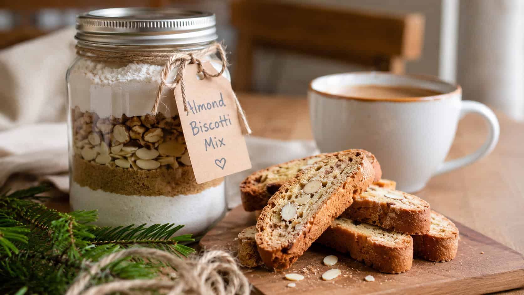 a mason jar of almond biscotti mix beside crisp sliced biscotti and a coffee cup in a wide horizontal shot, on a wooden table with twine and pine sprigs, natural light, shallow depth of field, landscape orientation