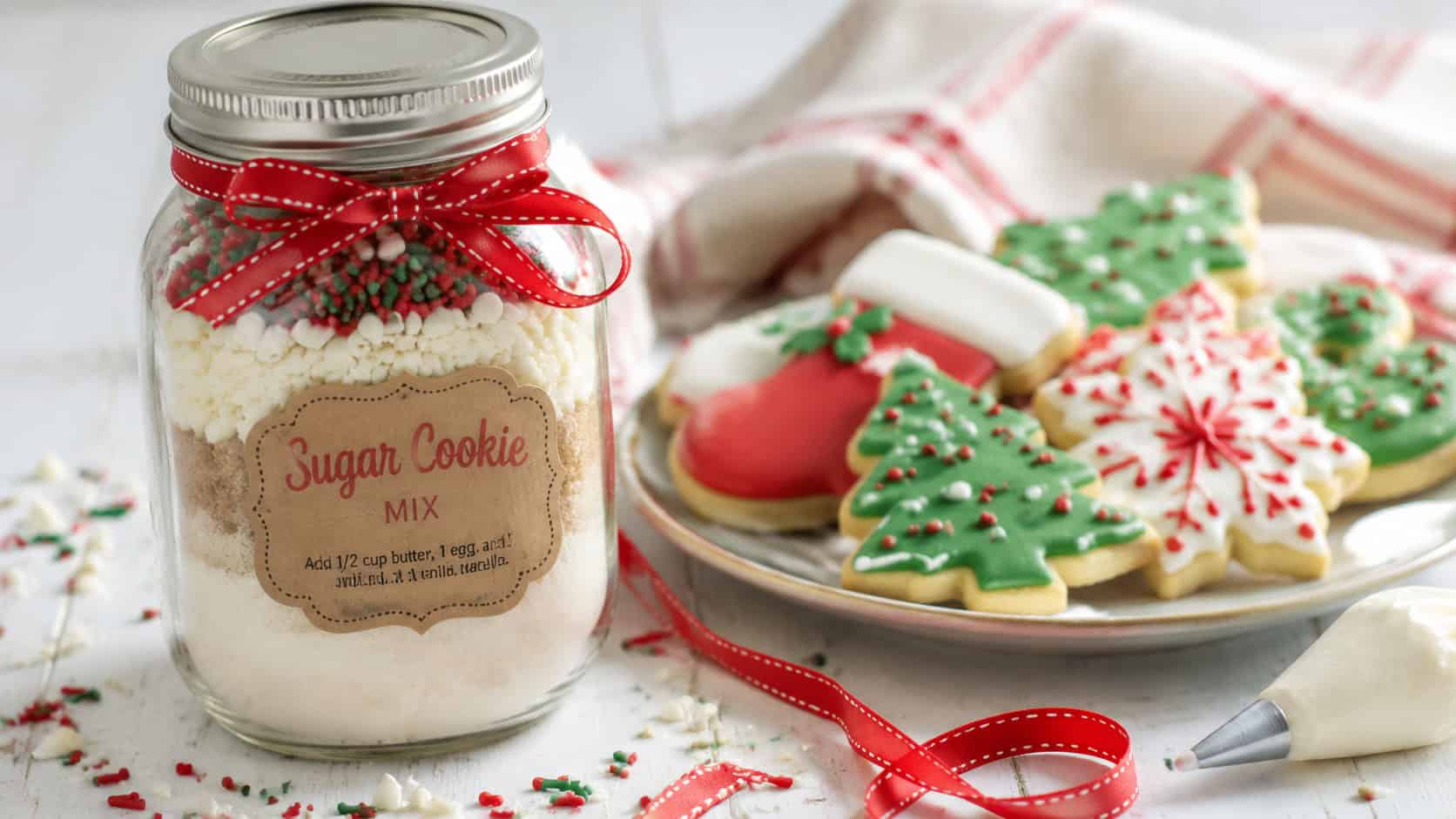 a mason jar of sugar cookie mix beside decorated cut-out holiday cookies in a wide horizontal shot, on a white surface with icing, sprinkles, and a red ribbon, natural light, shallow depth of field, landscape orientation