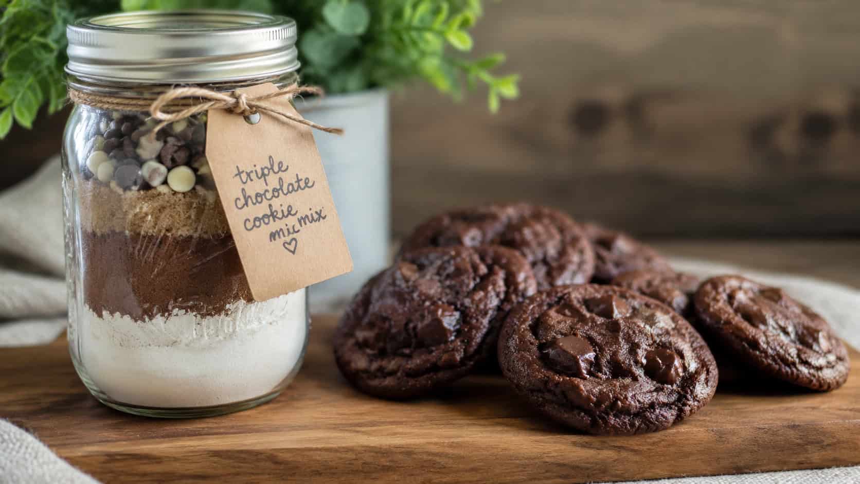 a mason jar of triple chocolate cookie mix beside fudgy chocolate chunk cookies in a wide horizontal shot, on a wooden board with a kraft gift tag and ribbon, natural light, shallow depth of field, landscape orientation