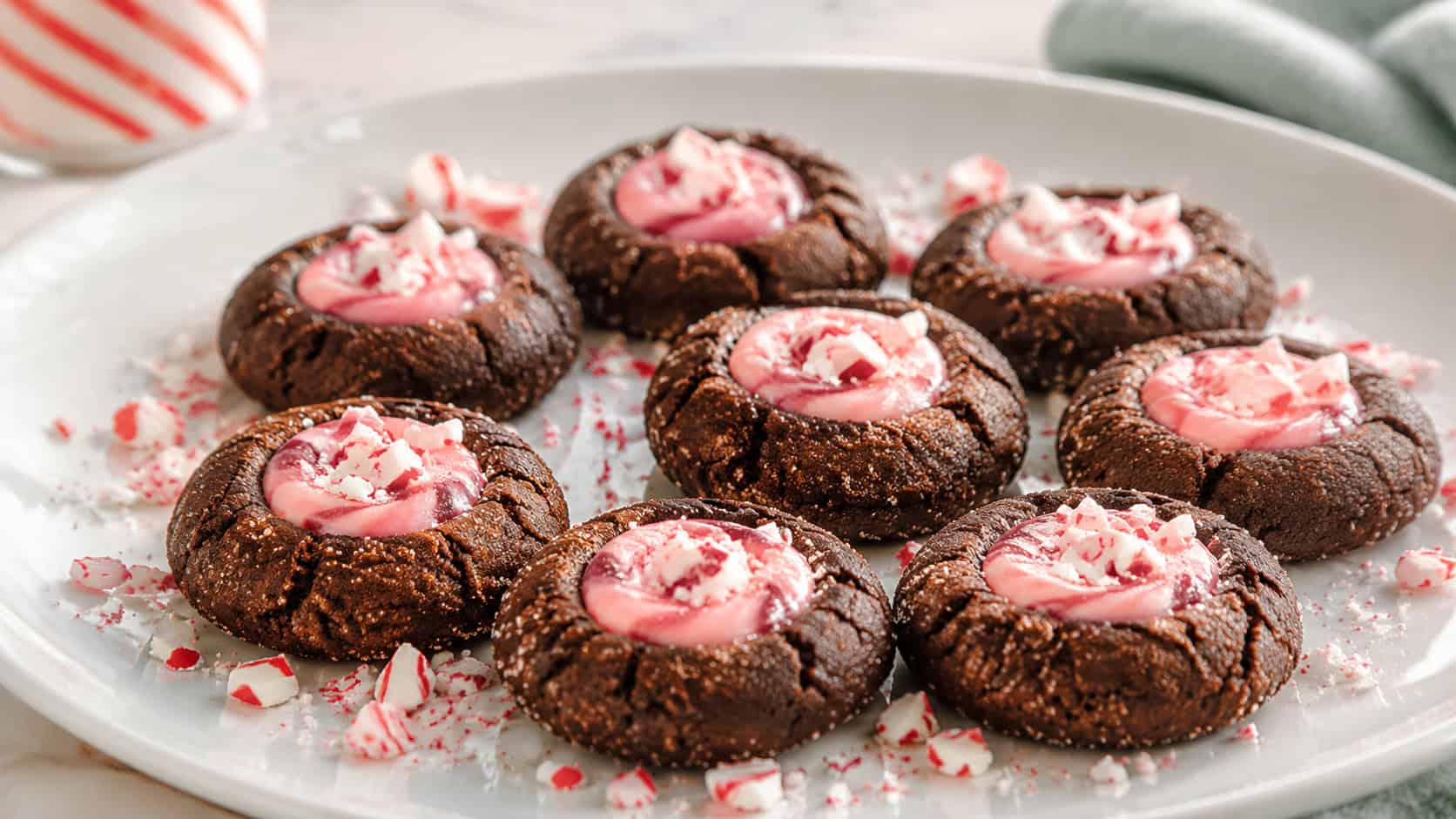 chocolate thumbprint cookies filled with glossy peppermint ganache arranged wide on a white plate, crushed candy cane bits scattered around, bright natural light, shallow depth of field, landscape orientation
