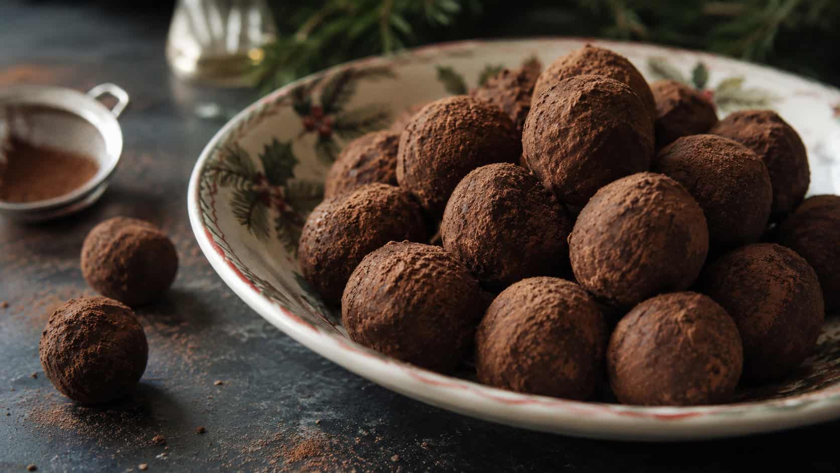 cocoa-dusted chocolate rum balls piled wide in a shallow festive bowl on a dark table, a few loose balls and cocoa nearby, moody natural light, shallow depth of field, horizontal landscape composition