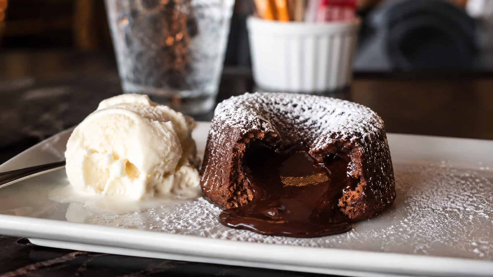 a chocolate lava cake cut open with molten center flowing onto a plate, dusted with powdered sugar and a scoop of vanilla ice cream beside it, shown wide across a dark table, warm natural light, shallow depth of field, horizontal landscape composition