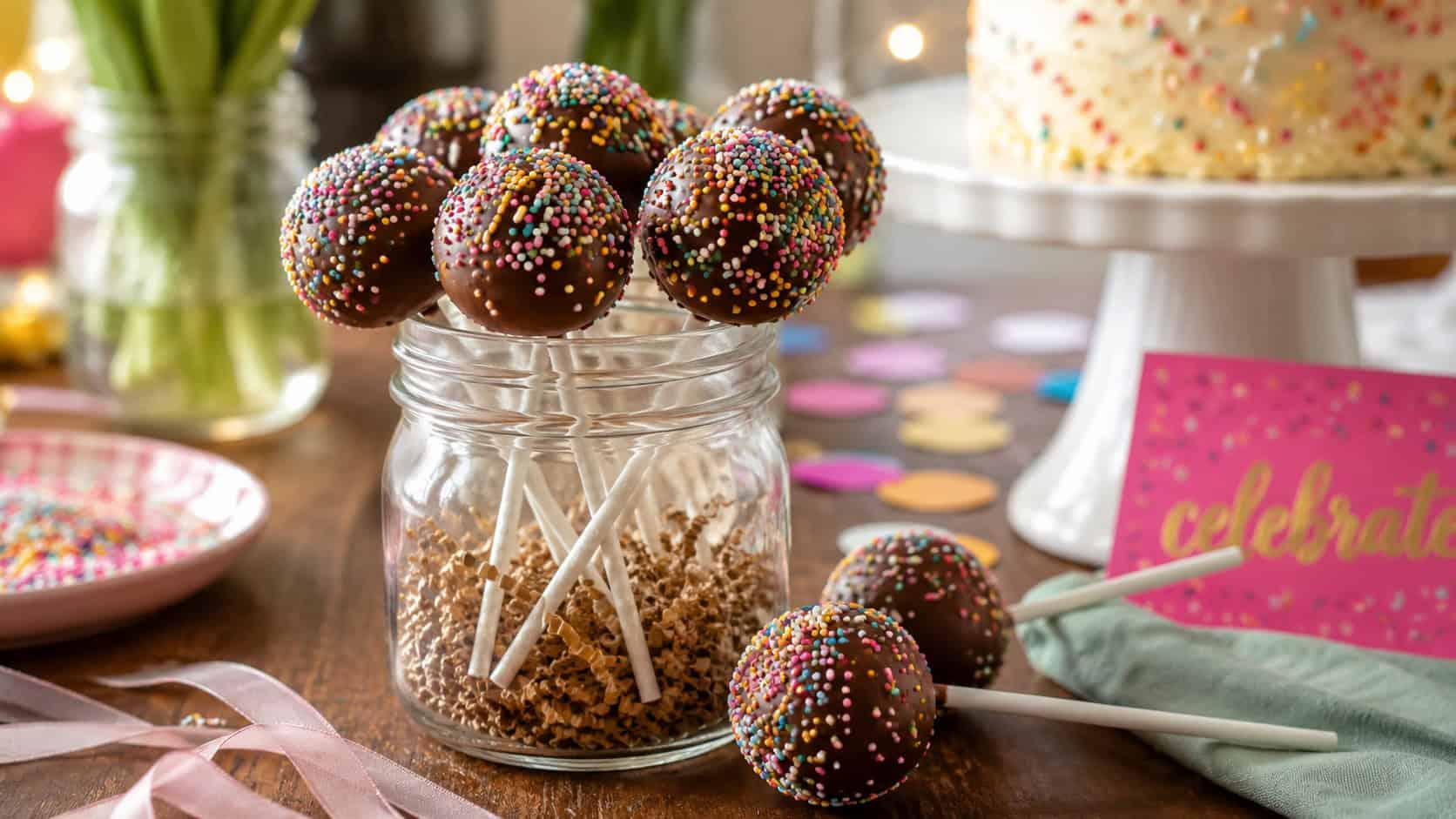 chocolate cake pops covered in sprinkles standing upright in a jar shown wide across a festive table, a few finished pops resting nearby, warm natural light, shallow depth of field, horizontal landscape composition