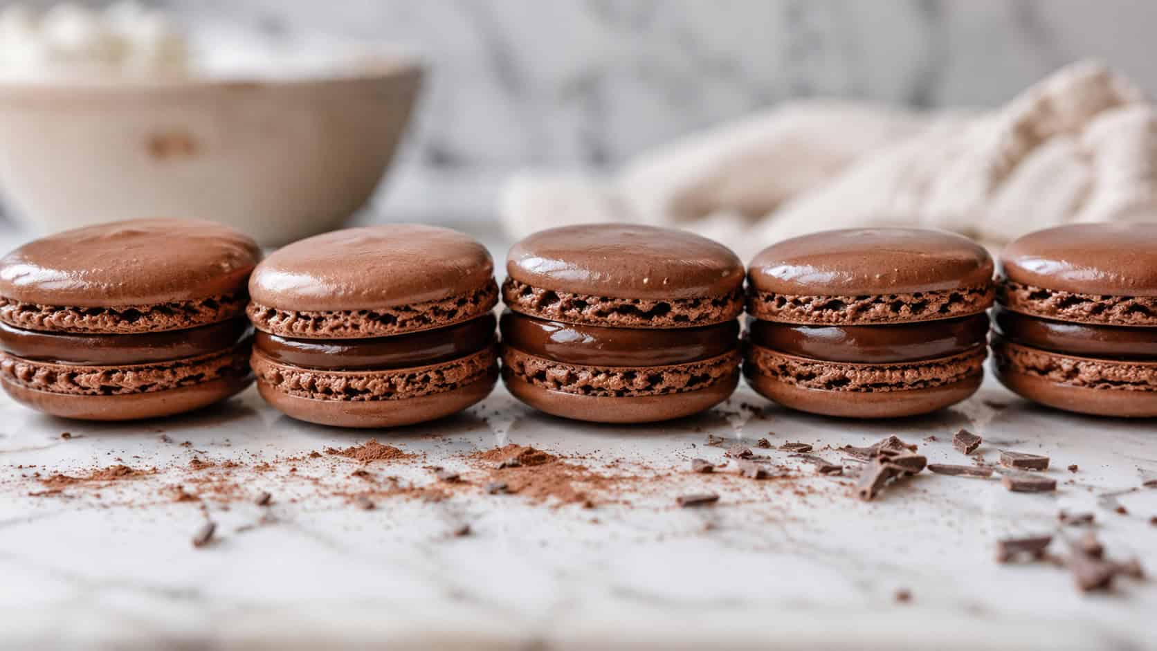 glossy chocolate macarons lined up wide across a white marble surface, a dusting of cocoa and a few chocolate shavings nearby, soft natural light, shallow depth of field, landscape orientation