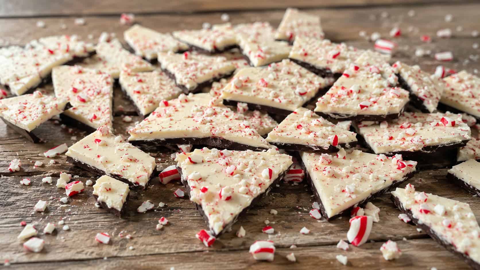 broken peppermint bark shards spread wide across a rustic wooden table, red and white candy cane bits scattered around, soft natural light, shallow depth of field, horizontal landscape composition