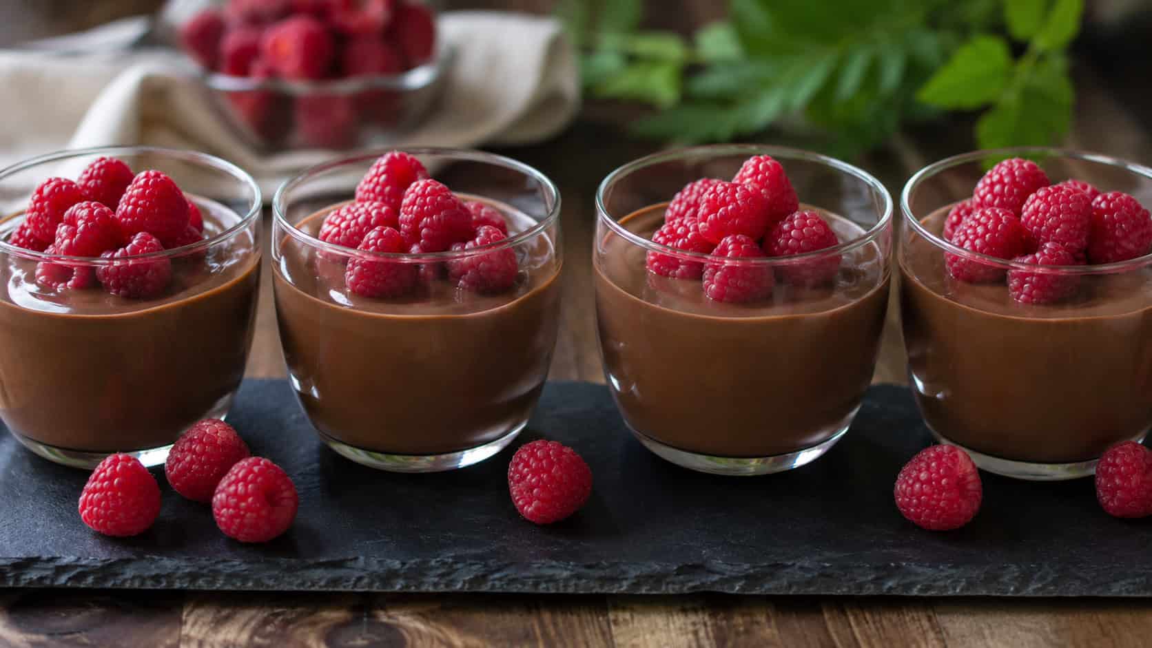 glossy chocolate panna cotta in glass cups topped with fresh raspberries lined up wide across a slate board, a few loose berries scattered around, soft natural light, shallow depth of field, horizontal landscape composition
