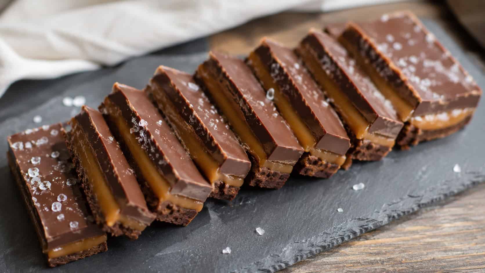 layered salted caramel chocolate bars cut and fanned wide across a slate board, glossy chocolate tops sprinkled with flaky salt, soft natural light, shallow depth of field, horizontal landscape composition