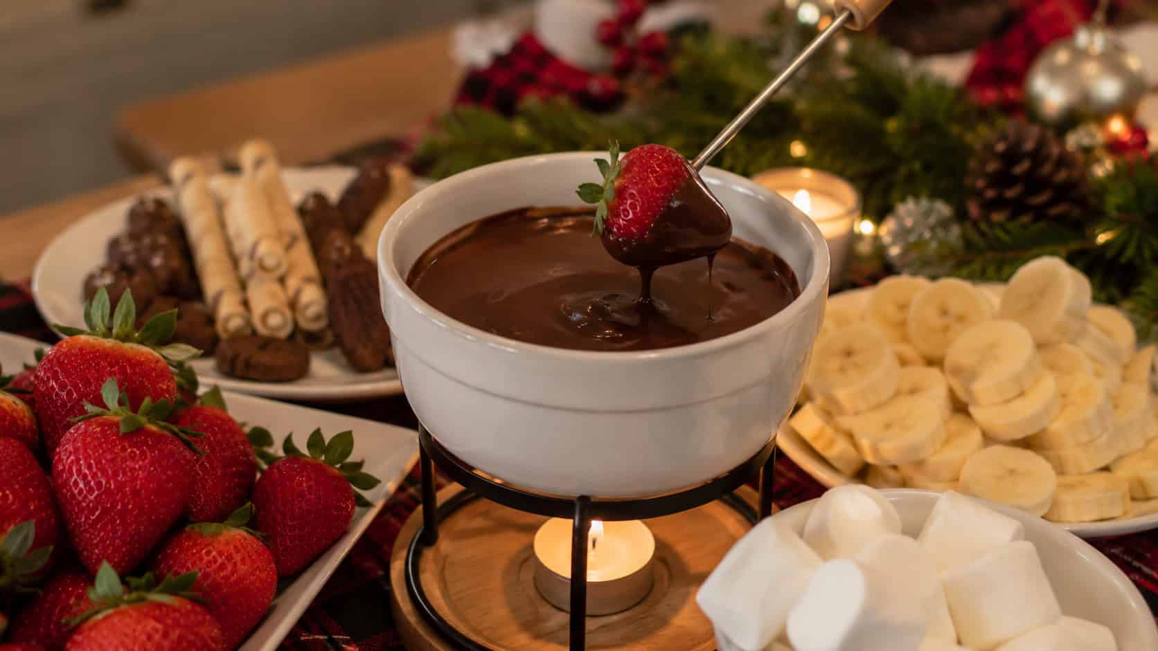 a warm pot of glossy melted chocolate fondue surrounded by strawberries, banana slices, and marshmallows arranged wide across a holiday table, soft candlelit natural light, shallow depth of field, horizontal landscape composition