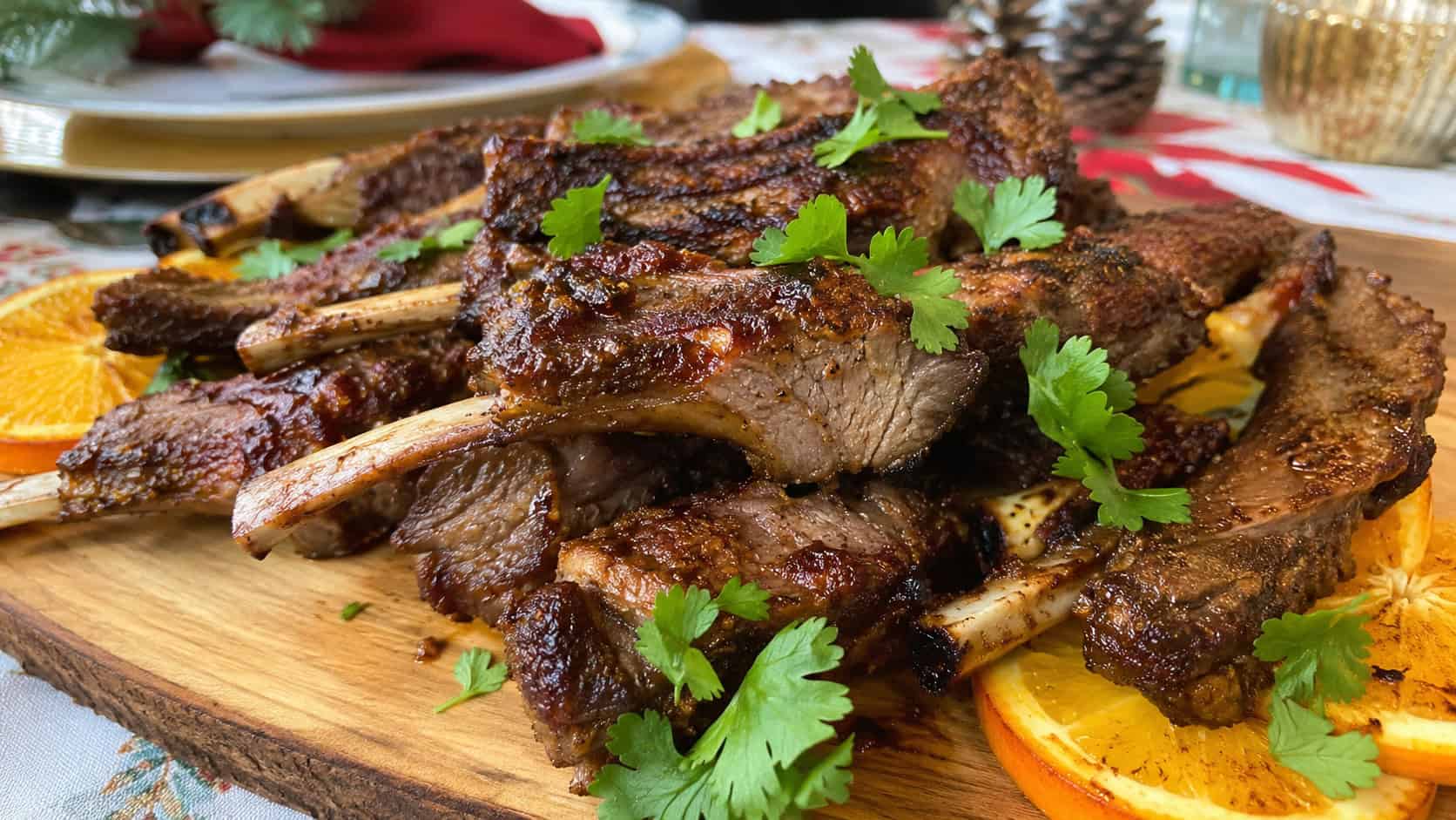 crisp-edged citrus spice-rubbed lamb ribs in a wide horizontal shot, piled on a wooden serving board with orange slices and a scatter of fresh cilantro on a holiday table, natural light, shallow depth of field, landscape orientation