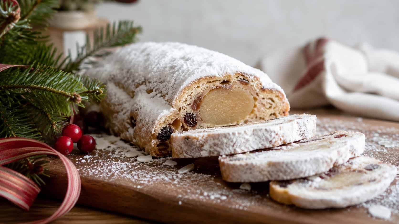 a sliced marzipan stollen loaf showing the almond center heavily dusted with powdered sugar arranged in a wide horizontal shot on a wooden board, holiday greenery and ribbon nearby, soft natural light, shallow depth of field, landscape orientation