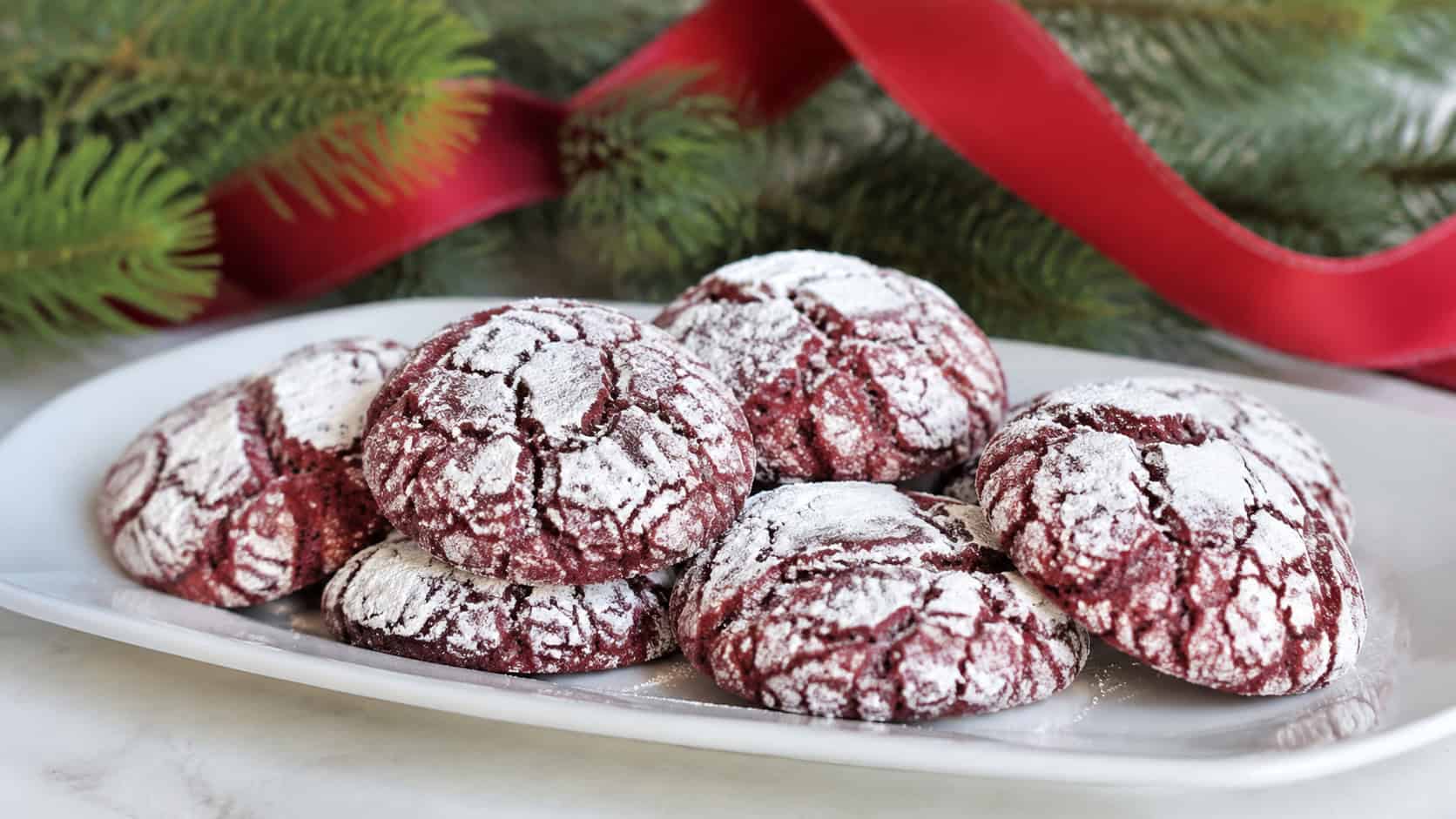 red velvet crinkle cookies with powdered sugar crackle tops arranged in a wide horizontal shot on a white plate, red ribbon and pine sprigs in the background, soft natural light, shallow depth of field, landscape orientation