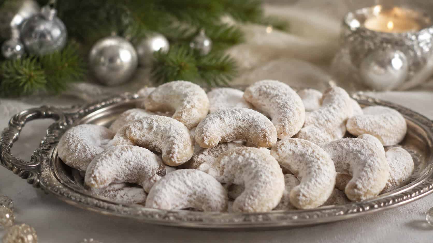 crescent-shaped almond cookies dusted with powdered sugar arranged in a wide horizontal shot on a vintage tray, evergreen and silver ornaments in the background, soft natural light, shallow depth of field, landscape orientation