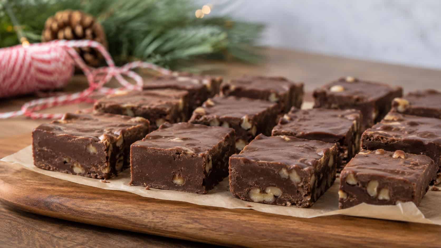 squares of rich chocolate walnut fudge arranged in a wide horizontal shot on a piece of parchment on a wooden board, holiday twine and pine in the background, soft natural light, shallow depth of field, landscape orientation