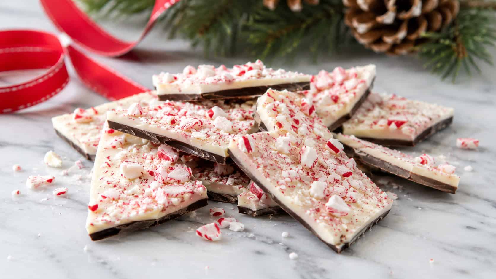broken peppermint bark shards with dark and white chocolate layers topped with crushed candy cane arranged in a wide horizontal shot on a white marble surface, red ribbon and pine in the background, soft natural light, shallow depth of field, landscape orientation