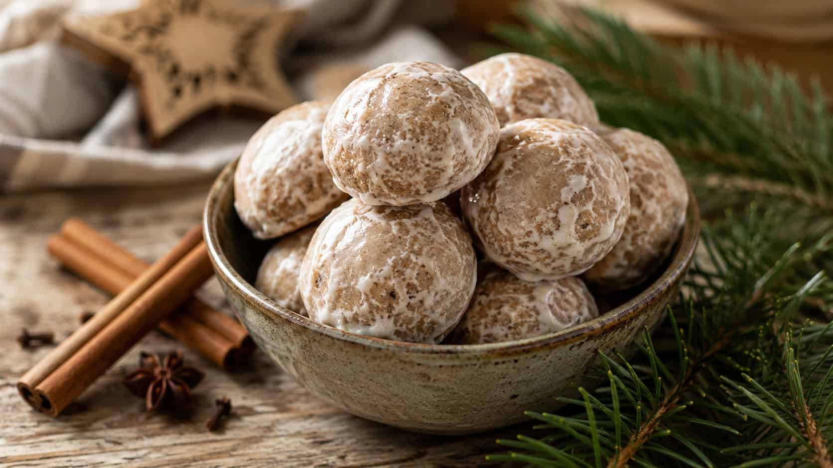 round glazed Pfeffernusse spice cookies arranged in a wide horizontal shot in a small bowl on a rustic table, cinnamon sticks and pine sprigs nearby, warm natural light, shallow depth of field, landscape orientation