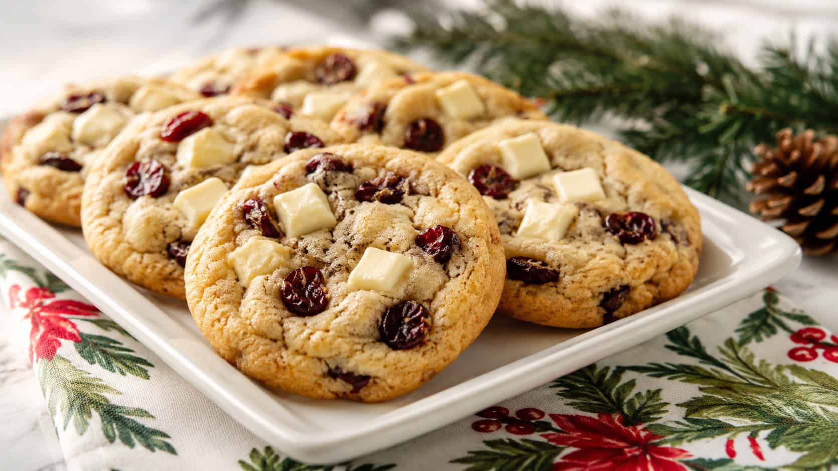 soft cookies dotted with white chocolate chunks and red cranberries arranged in a wide horizontal shot on a white plate, holiday napkin and pine sprigs nearby, bright natural light, shallow depth of field, landscape orientation
