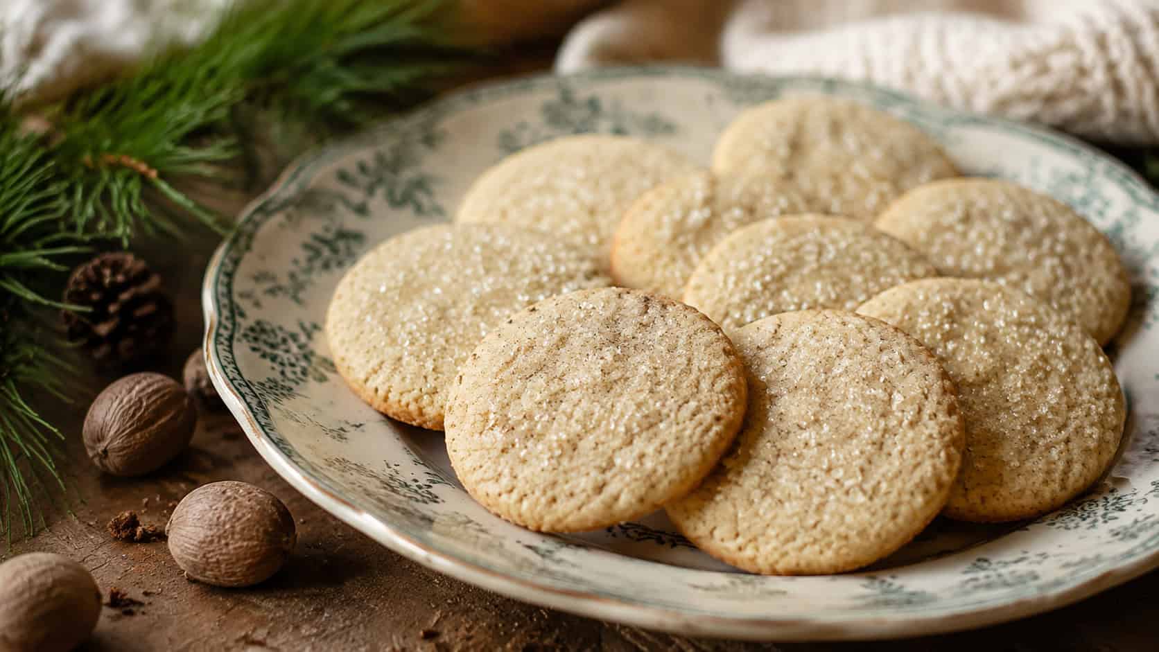 round nutmeg sugar cookies topped with sparkling sugar arranged in a wide horizontal shot on a vintage plate, nutmeg and pine on a cozy table, soft natural light, shallow depth of field, landscape orientation