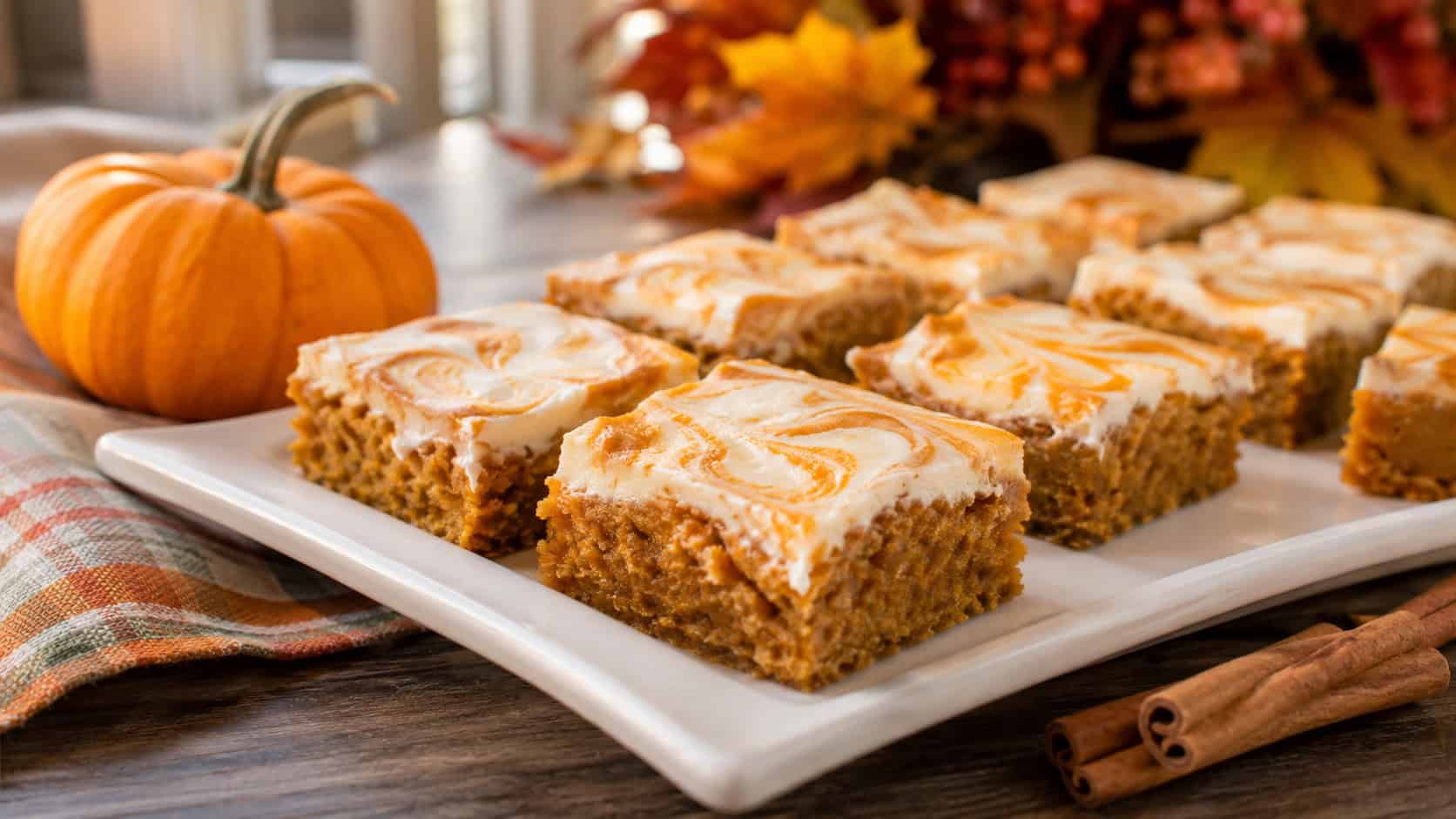 pumpkin spice bars with swirled cream cheese frosting arranged in a wide horizontal shot on a white platter, cinnamon sticks and a small pumpkin nearby, warm natural light, shallow depth of field, landscape orientation