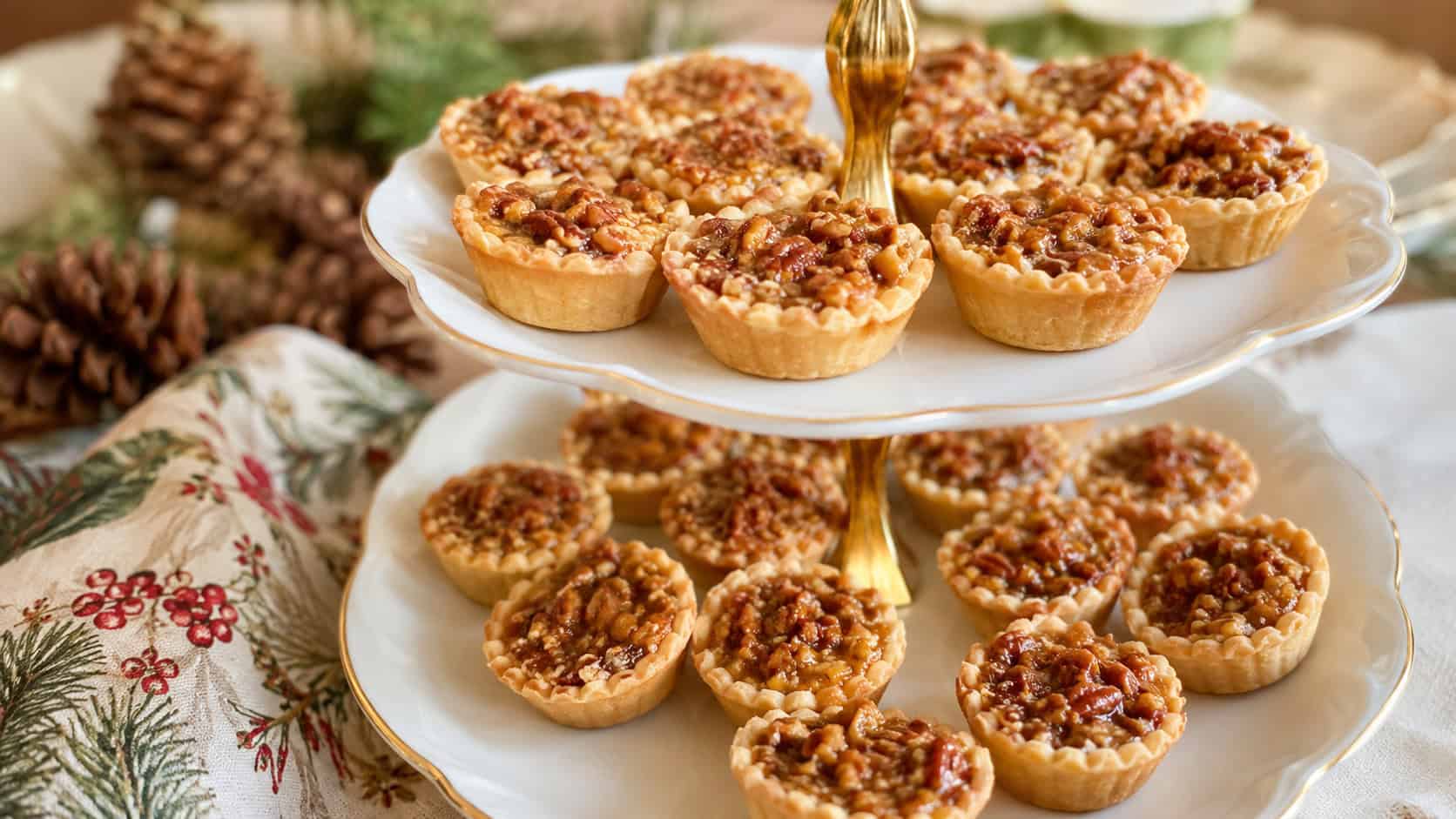 mini pecan tassies with golden buttery filling arranged in a wide horizontal shot on a tiered serving plate, festive napkin and pinecones in the background, warm natural light, shallow depth of field, landscape orientation