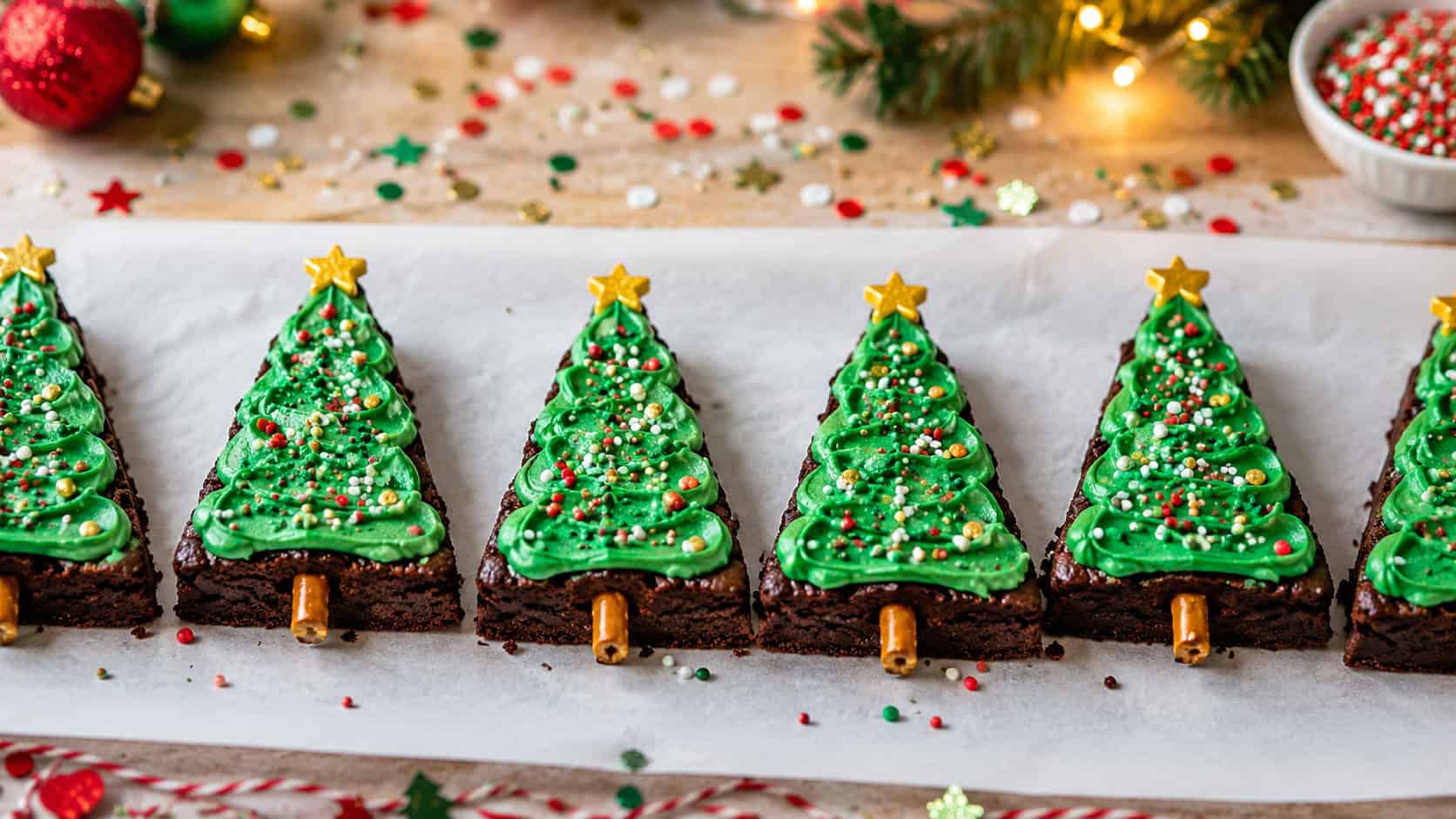 Christmas tree shaped brownies lined up on parchment in a wide horizontal shot, fudgy triangles frosted green with colorful sprinkle ornaments and a star on top, festive table scattered with confetti, bright cheerful light, shallow depth of field, landscape orientation