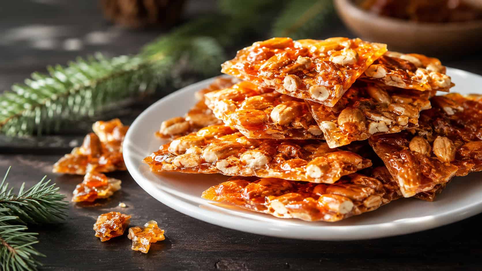 golden nut brittle shards stacked on a white plate in a wide horizontal shot, glossy amber candy packed with roasted nuts, a few broken pieces and pine sprigs on a dark table, dramatic side light, shallow depth of field, landscape orientation