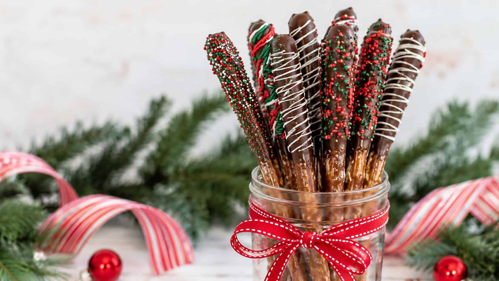 chocolate-dipped pretzel rods standing in a tall mason jar in a wide horizontal shot, dark chocolate ends covered in red and green sprinkles with white drizzle, festive ribbon and pine nearby, bright cheerful light, shallow depth of field, landscape orientation
