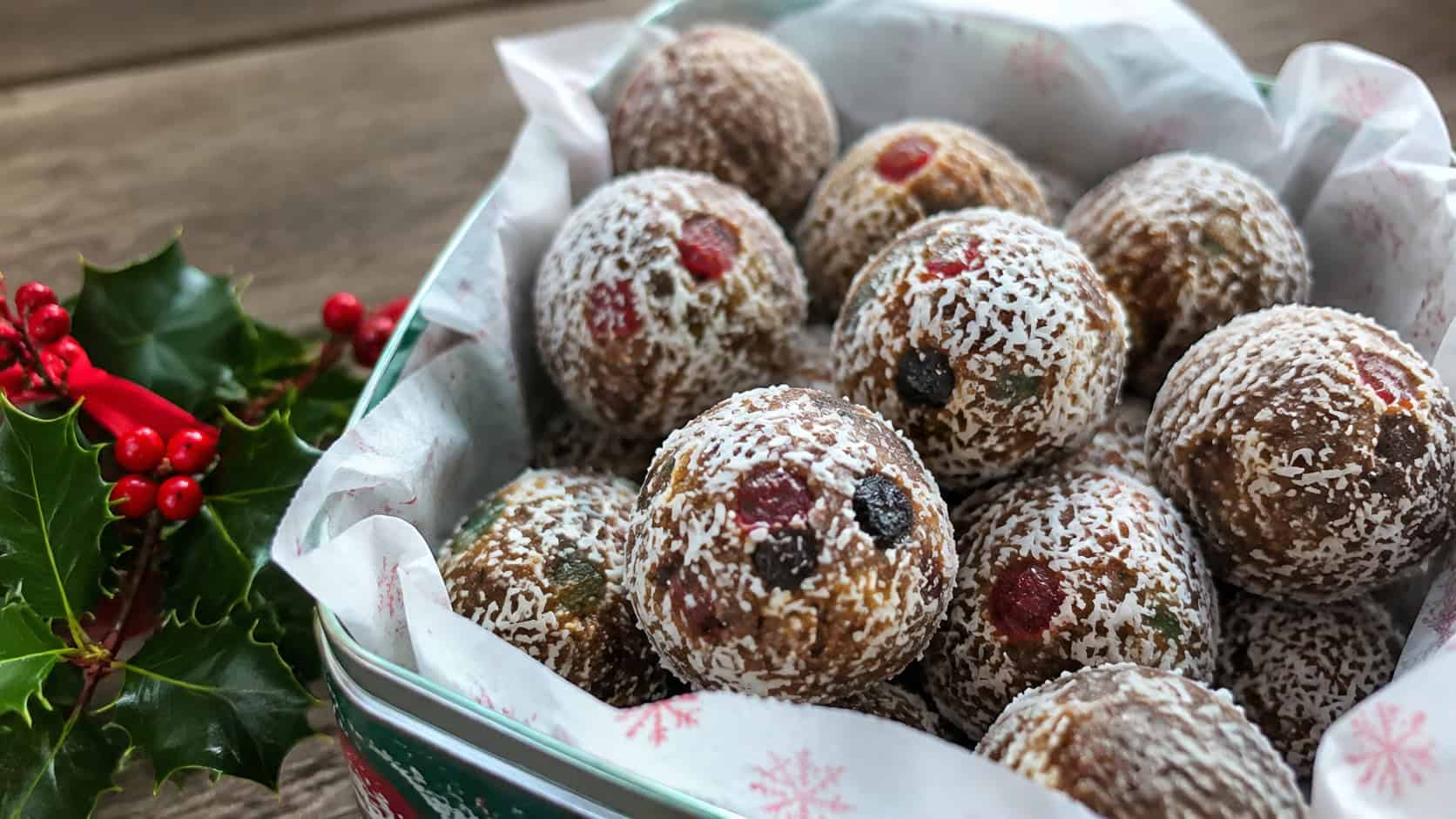 coconut-coated fruitcake bites arranged in a paper-lined tin in a wide horizontal shot, dense fruit and nut balls dusted in white coconut, a sprig of holly and red berries beside them, soft natural light, shallow depth of field, landscape orientation