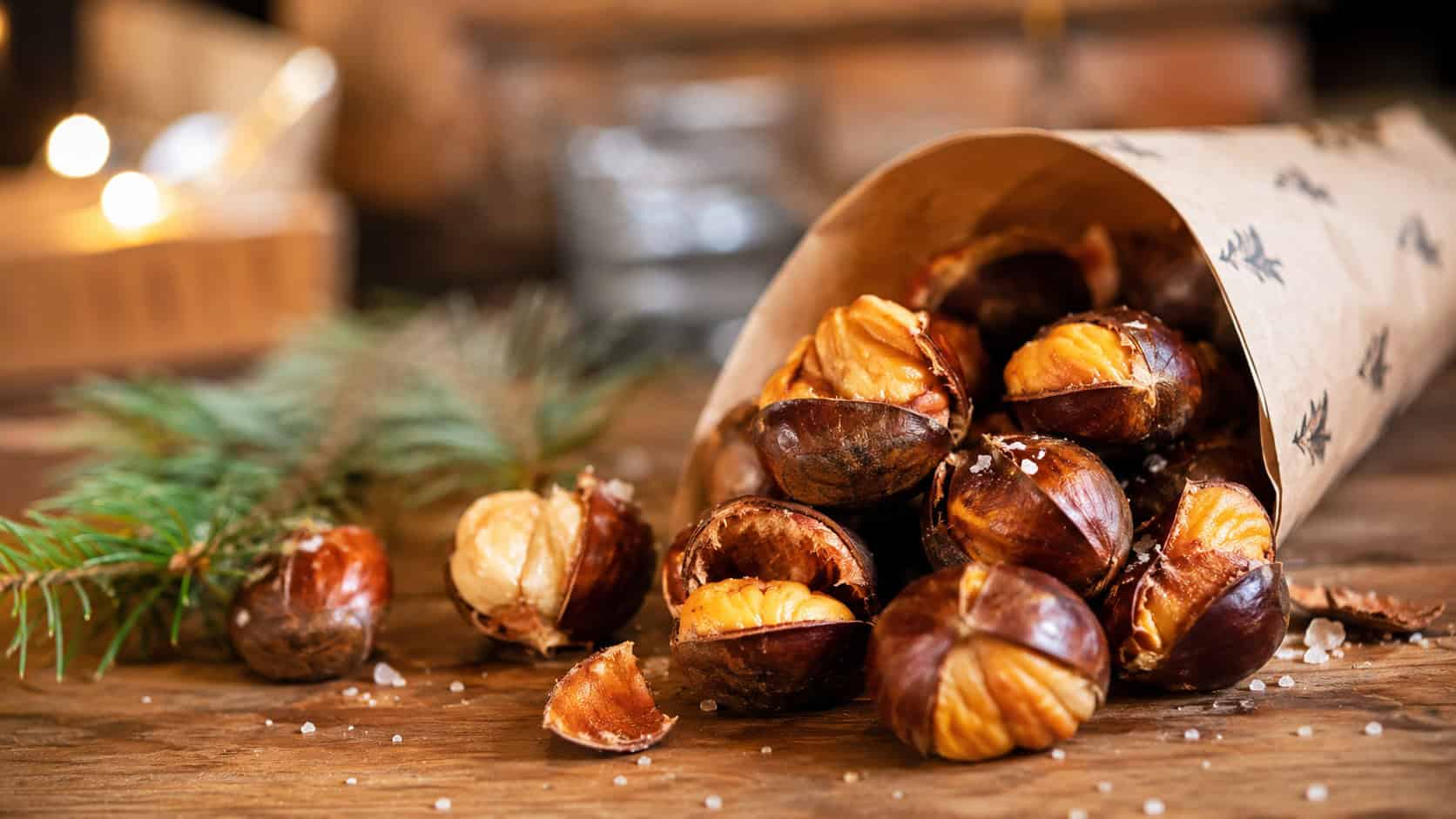 warm roasted chestnuts spilling from a paper cone onto a wooden table in a wide horizontal shot, split caramel-brown shells and creamy centers, a sprinkle of sea salt and pine sprigs nearby, cozy golden light, shallow depth of field, landscape orientation
