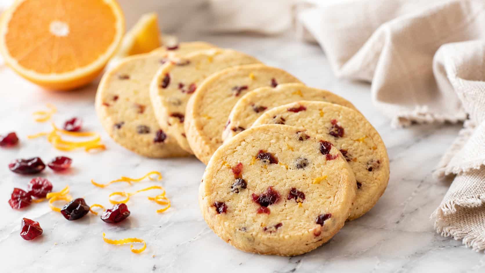 cranberry orange shortbread rounds fanned across a marble surface in a wide horizontal shot, pale golden cookies flecked with red cranberries, curls of orange zest and a linen napkin beside them, bright airy light, shallow depth of field, landscape orientation