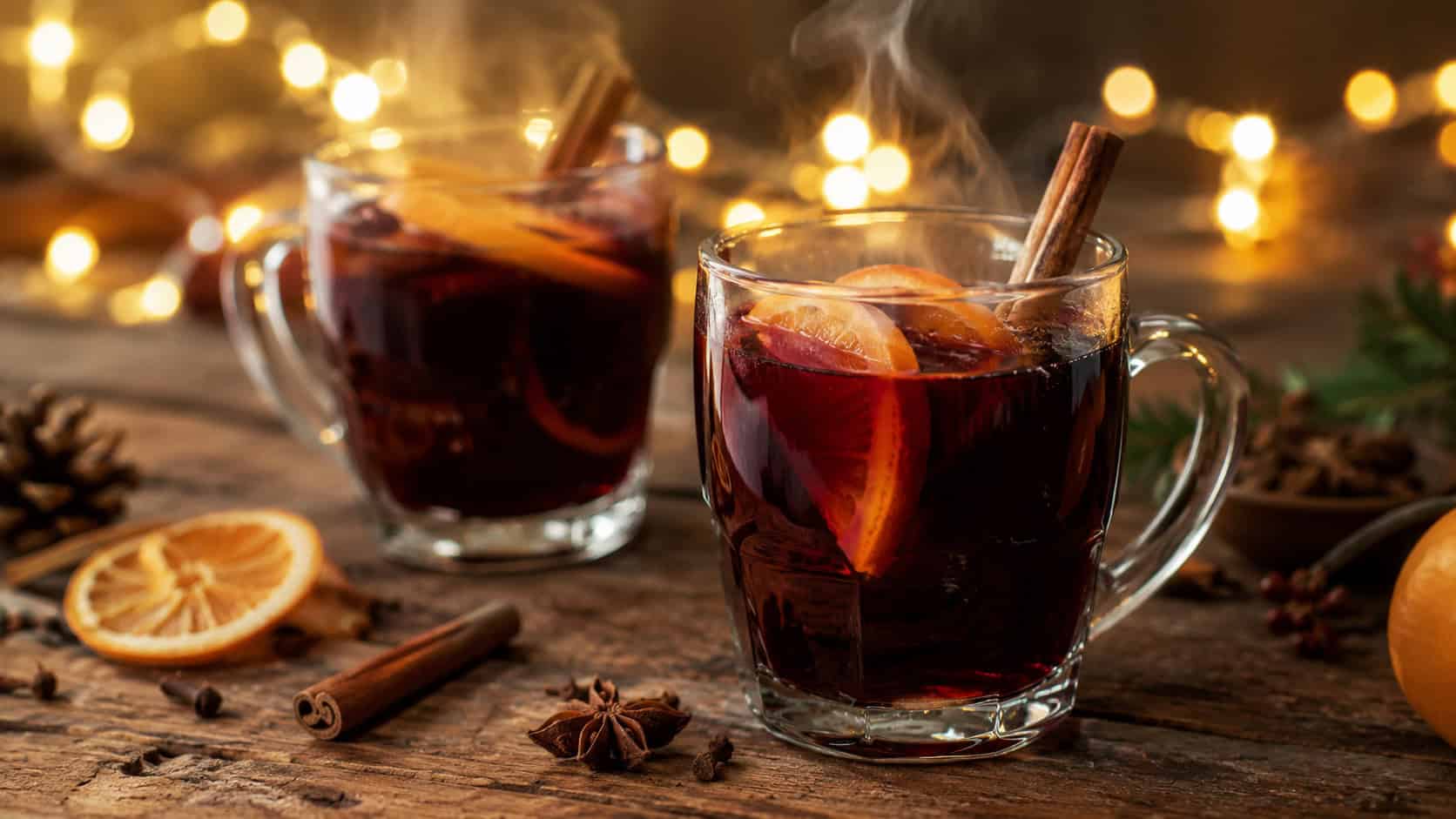steaming mugs of spiced mulled red wine with orange slices and cinnamon sticks, in a wide horizontal shot on a wooden table with string lights glowing softly in the background, warm cozy natural light, shallow depth of field, landscape orientation