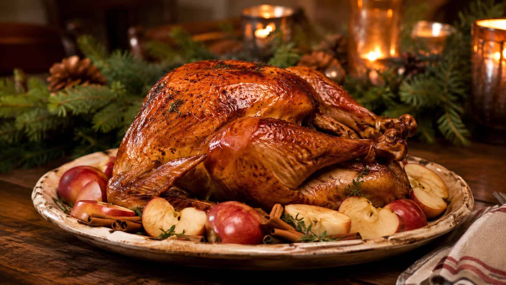 an apple cider brined turkey on a rustic platter in a wide horizontal shot, burnished golden skin surrounded by apple halves and cinnamon sticks, pine garland and candles behind, warm cozy light, shallow depth of field, landscape orientation