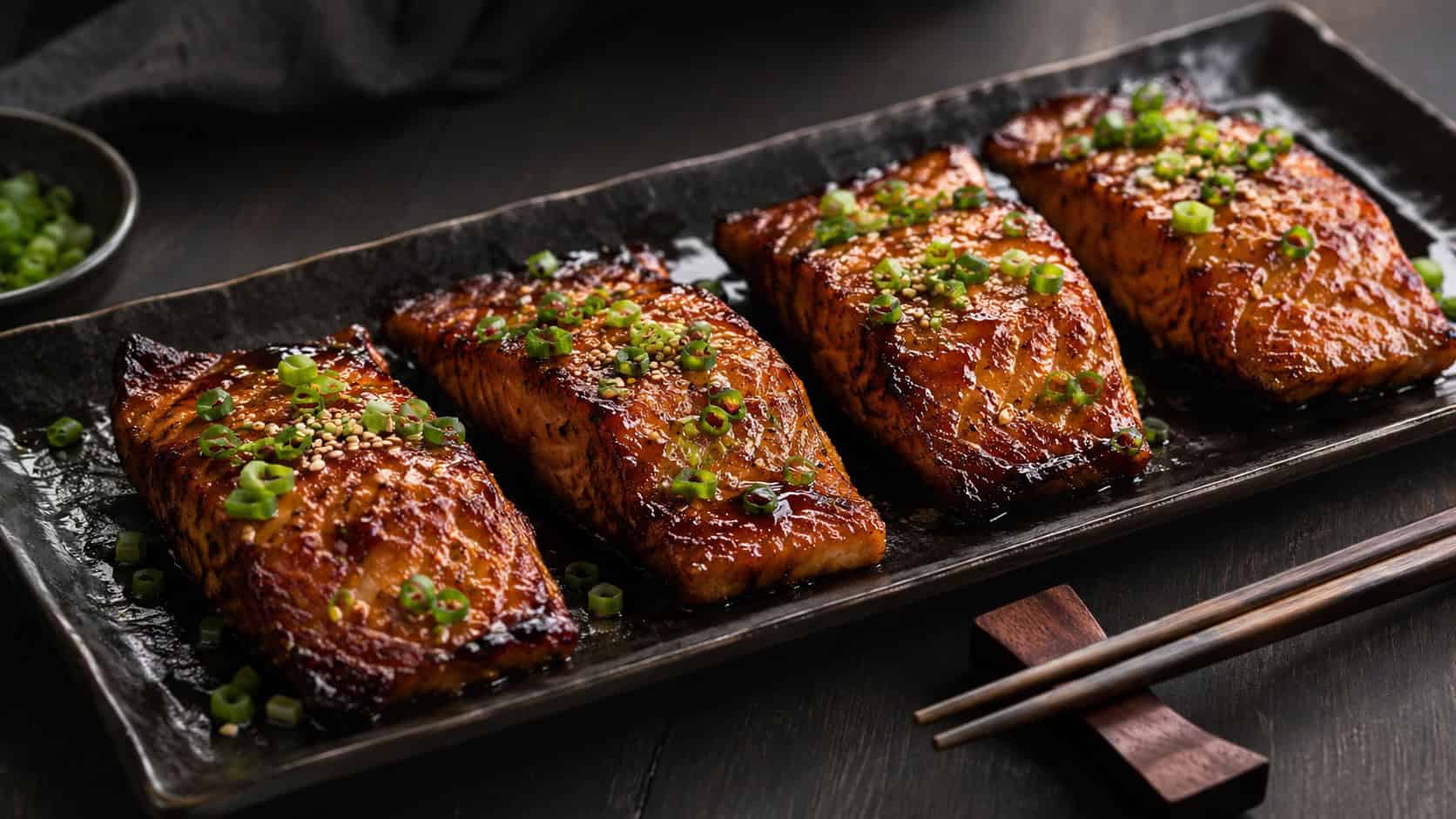 four glossy ginger-soy glazed trout fillets with caramelized edges, scattered with green onion and sesame seeds, arranged in a wide horizontal shot on a black rectangular plate with chopsticks beside, on a dark table, moody natural light, shallow depth of field, landscape orientation