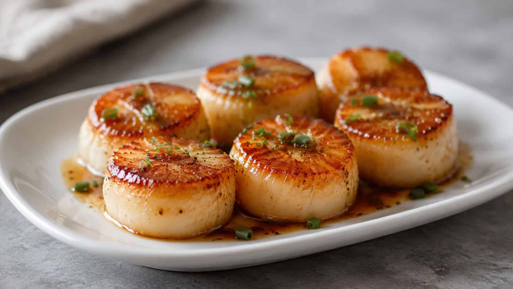 plump golden-seared scallops glazed and glistening, arranged in a wide horizontal shot on a small white plate with a drizzle of glaze and a sprinkle of chives, on a soft gray surface, elegant natural light, shallow depth of field, landscape orientation