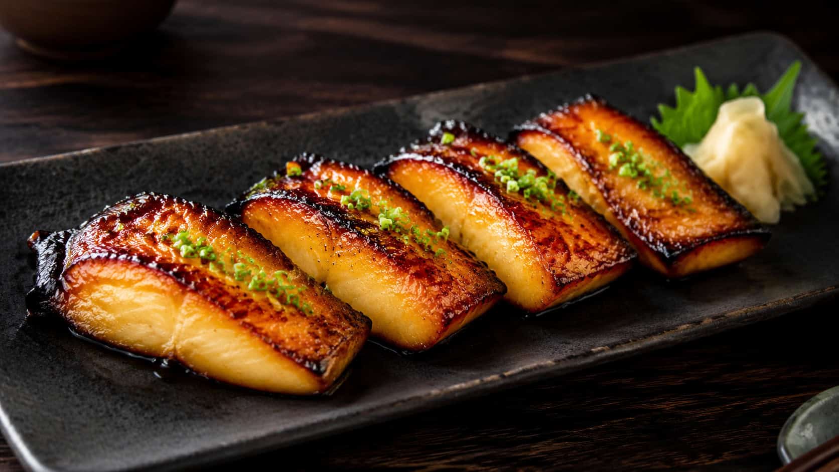 four lacquered miso-glazed black cod fillets with deeply caramelized golden-brown tops, plated in a wide horizontal shot on a matte black dish with pickled ginger and green onion, on a dark wooden surface, dramatic natural light, shallow depth of field, landscape orientation