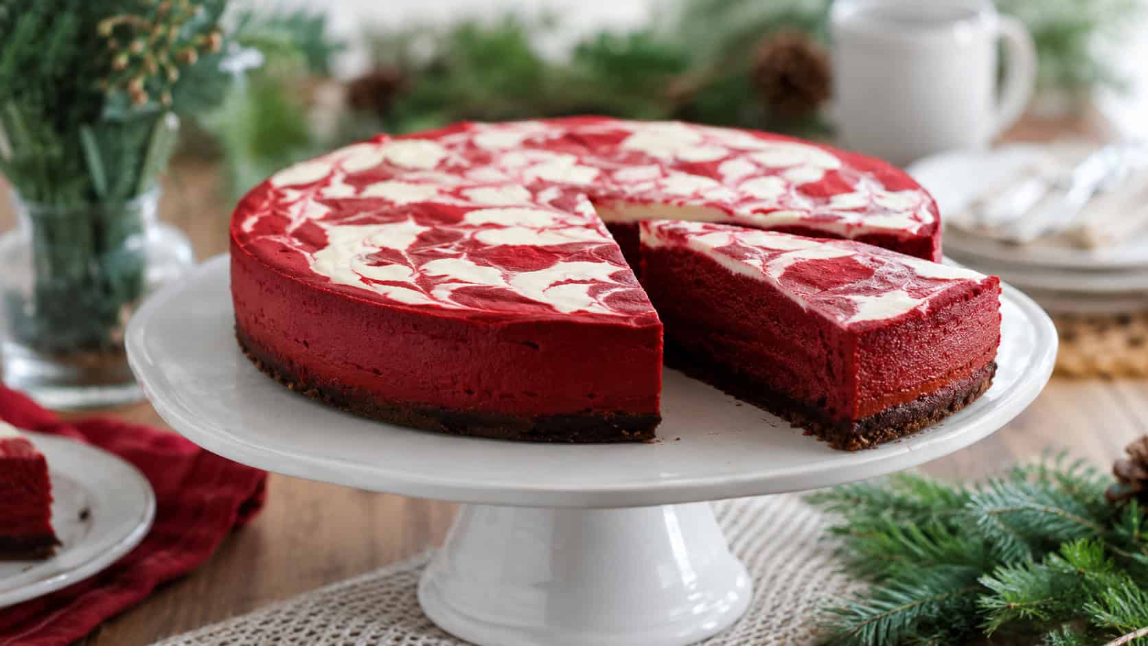 a red velvet cheesecake on a white cake stand in a wide horizontal shot, deep crimson layer with white cream cheese swirl on top, one slice cut, on a festive table with greenery, soft natural light, shallow depth of field, landscape orientation