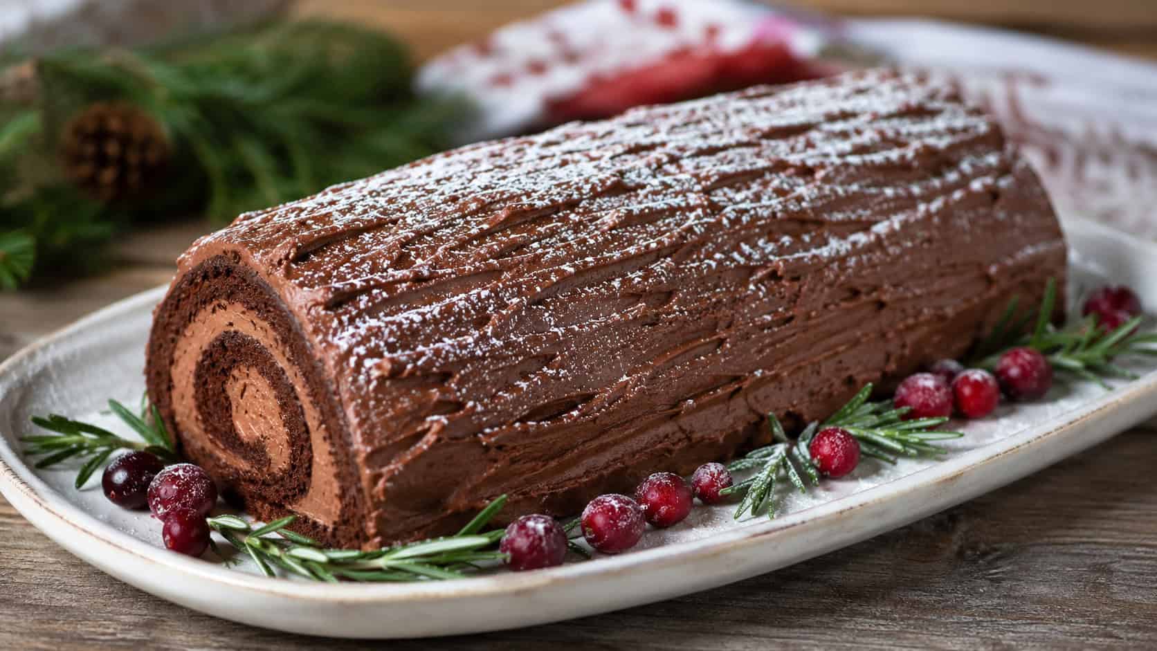 a chocolate yule log cake with bark-textured frosting and a dusting of powdered sugar snow in a wide horizontal shot, on a festive platter with rosemary and cranberries, natural light, shallow depth of field, landscape orientation