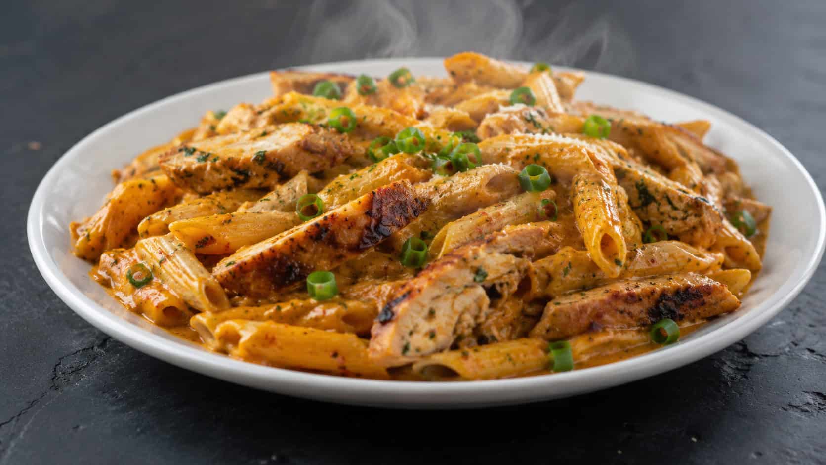 a steaming plate of spicy creamy Cajun penne with a reddish-orange sauce and charred chicken pieces, sprinkled with green onion, in a wide horizontal shot on a dark slate surface, natural light, shallow depth of field, landscape orientation