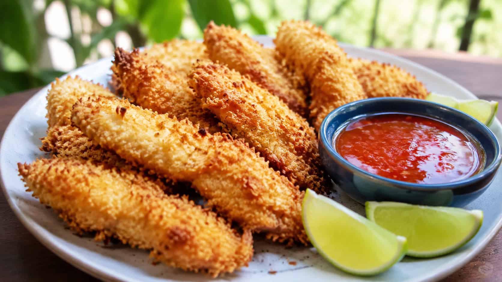 golden coconut-crusted chicken strips in a wide horizontal shot, arranged on a plate with a dish of sweet chili sauce and lime wedges, natural light, shallow depth of field, landscape orientation