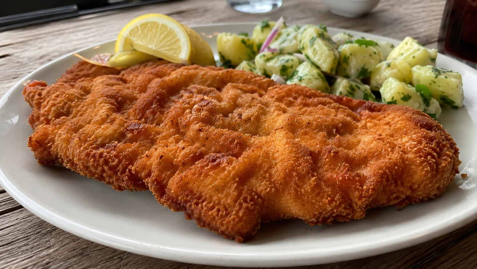 a large golden crispy chicken schnitzel in a wide horizontal shot, plated with lemon wedges and a side of potato salad, natural light, shallow depth of field, landscape orientation
