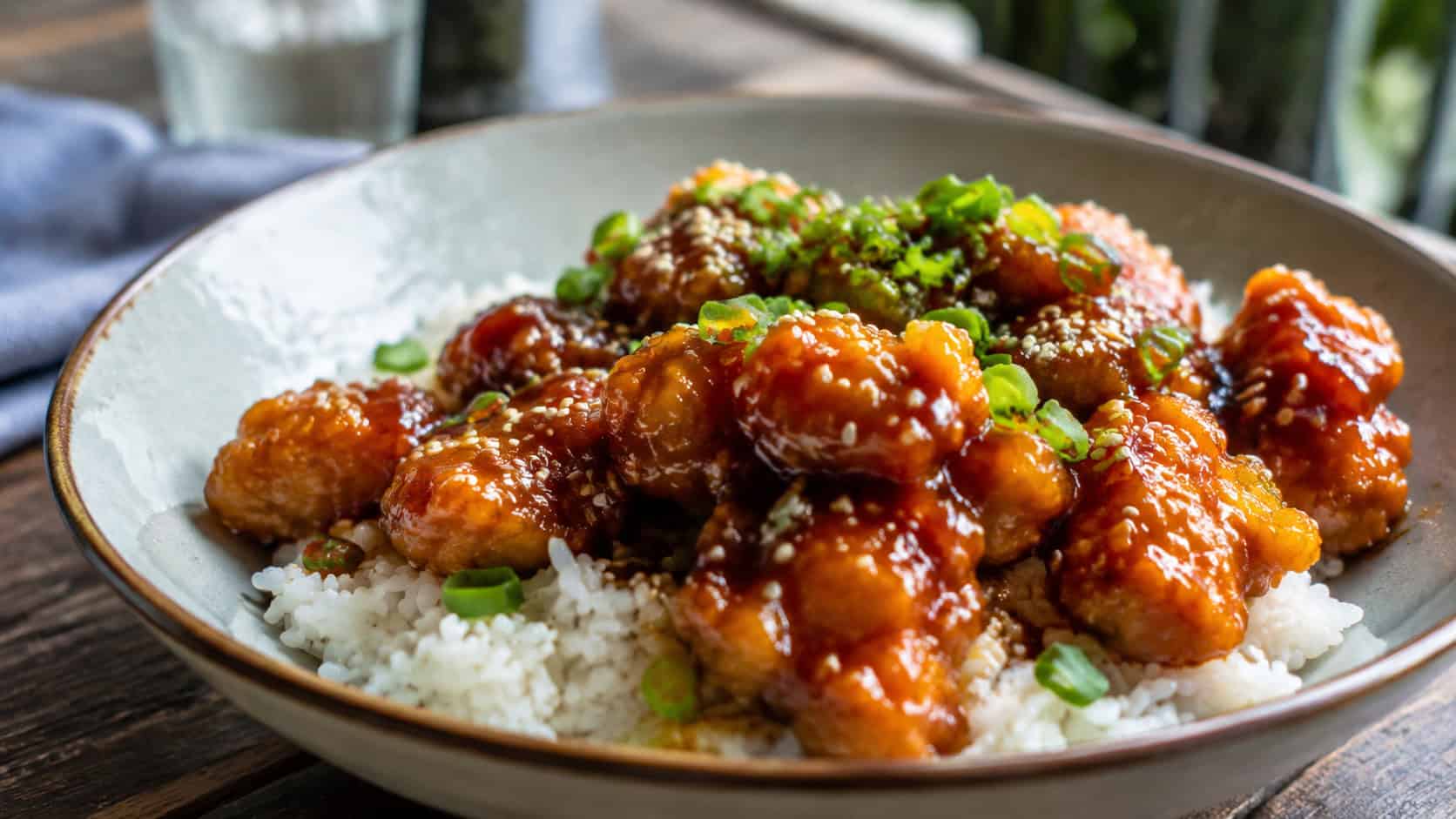 glossy honey garlic breaded chicken bites in a wide horizontal shot, served over white rice with sesame seeds and green onion in a shallow bowl, natural light, shallow depth of field, landscape orientation