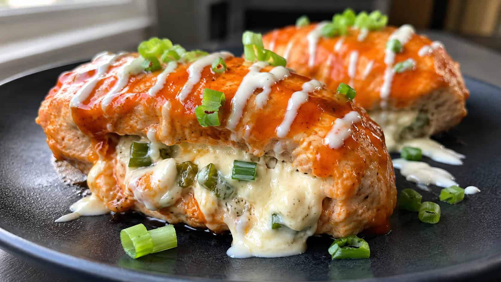 buffalo chicken breasts glazed in orange sauce with creamy jalapeño filling oozing out, on a dark plate, wide horizontal shot, ranch drizzle and green onion garnish, natural light, shallow depth of field, landscape orientation